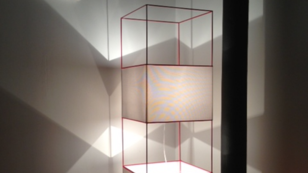 Lampadaire à structure en métal rouge et abat-jour blanc, composé de cubes superposés, projetant un jeu d’ombres graphiques sur le mur